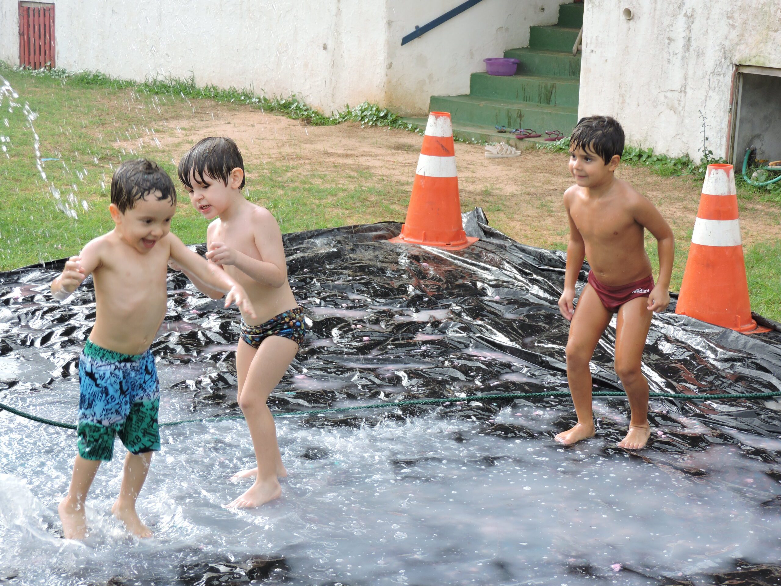 Três crianças alegres brincam de banho de mangueira durante Colônia de Férias da Casa Escola